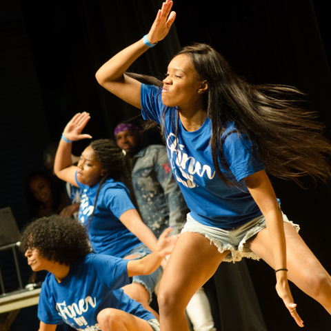 A group of students perform energetic dance moves on stage during the NPHC Stroll Off at Indiana State University, wearing matching blue shirts and shorts.