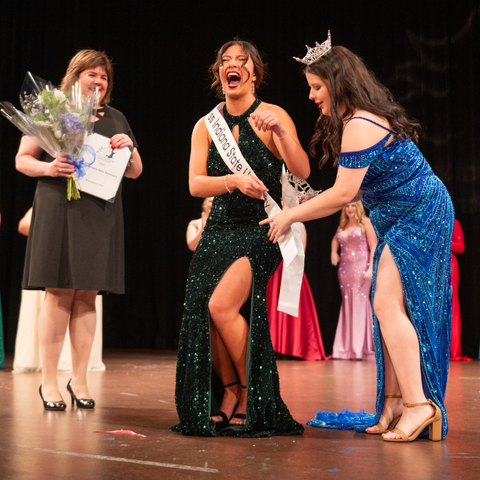 A pageant winner, wearing a sparkling green gown, reacts with joy as she is crowned by the previous titleholder during an award ceremony on stage. A bouquet of flowers is held by an official nearby