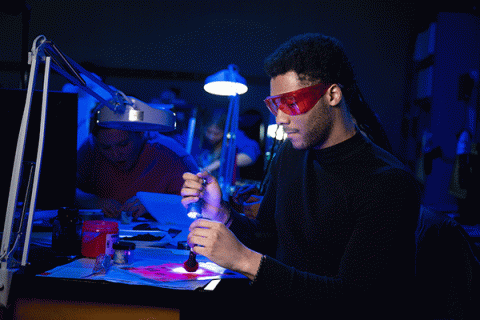 A Black male student wearing red glasses uses a flashlight to illuminate evidence in a crime lab lit by fluorescent black lights and a bright white lamp in the background. 