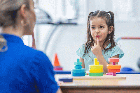 A young child, a girl with brown hair, sits at a table with colorful blocks visible on the table. She wears a light blue T-shirt. She presses her index finger against her chin. Seated opposite her, in the foreground and out of focus, is a woman with blond hair. She wears a blue T-shirt. The background is blurred and an orange cone is visible behind the girl to the left.