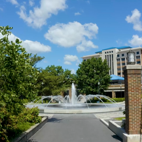 View of a campus walkway leading to a circular fountain surrounded by lush greenery, with a modern multi-story building in the background under a clear blue sky with scattered clouds.