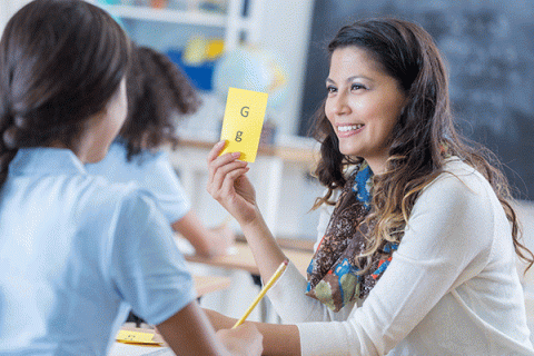 - A smiling woman with dark, medium-length hair and blonde highlights shows a yellow card with the letters ‘G’ and ‘g’ printed on it to a female student seated in front of her. The student has braided black hair and wears a blue shirt. She holds a pencil in her hand. The teacher wears a white long-sleeved top with a multi-colored scarf around her neck. Other students are visible in the background.