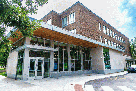 Exterior view of Dreiser Hall, a modern brick-and-glass building with large windows and a wooden overhang at the entrance. The entrance features double glass doors with the name 'Dreiser Hall' above them. There are trees and a car visible on the side, with a bright sky in the background.