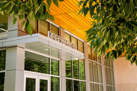 Exterior of a building with green leaves from a tree in the top of the frame in the foreground. The building’s entryway has a stained wood ceiling and a glass door and windows. A sign with what looks like silver metal letters sits above the entryway and reads “Dreiser Hall.”  