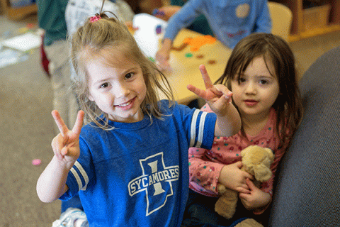 A young blonde girl wearing an Indiana State University shirt smiles at the camera in the Early Childhood Learning Center as a young dark-haired girl beside her looks on. Other children are partially visible around a table in the blurred background.