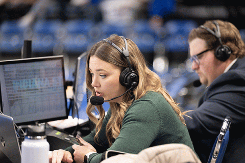 A female student with blondish-brown hair and wearing a green sweater is seated in a sports stadium, with rows of blue seats visibly blurred behind her. She is wearing headphones and a mic, holds a pen in her hand, and is looking at laptop and desktop computer monitors that are set up in front of her. A male student with brown hair and glasses, who is wearing a grey suit jacket and white collared shirt, is slightly blurred but visible behind her. He is wearing headphones.