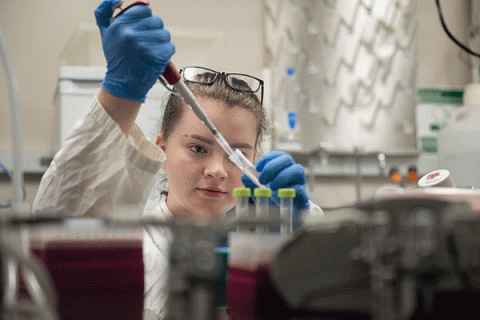 A female student with dark blonde hair, wearing a white lab coat, and with brown glasses pushed up onto her head, faces the camera as she uses a pipette in a lab setting. Various beakers and pieces of equipment are blurred but visible on a shelf in the foreground. 