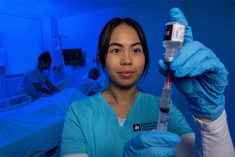 Tan-skinned female student with dark black hair and wearing blue scrubs looks closely at a hypodermic needle while drawing an injection, with another nurse working on a patient simulator in a hospital bed in the background. The room has a blue tint.   