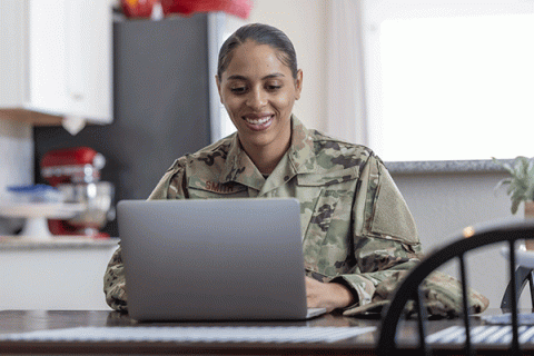 A Black woman wearing military fatigues sits and smiles in front of a laptop at a kitchen table in a residence.