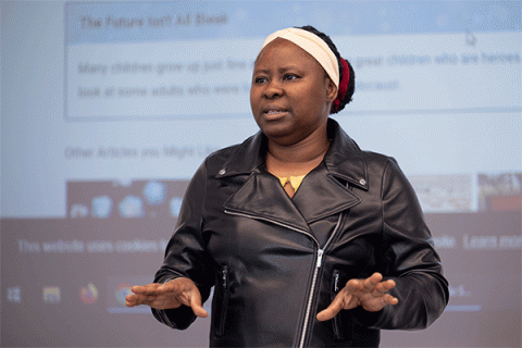 Black female professor wearing a black leather jacket and a white headscarf stands in front of an illuminated screen displaying a web page as she speaks to a class.