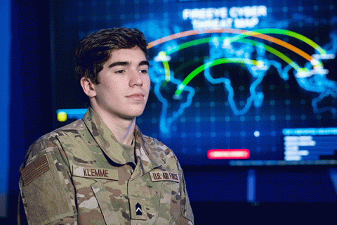 Male student with short, dark hair, and wearing military fatigues with an ‘Air Force’ patch, stands looking left to right in a darkly lit classroom with a bright, digital world map visible in the background. 