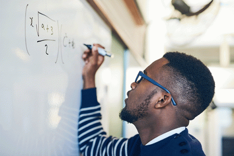 A Black man wearing glasses and a blue-and-white striped sweater writes a mathematical equation on a white board in a classroom or office setting.