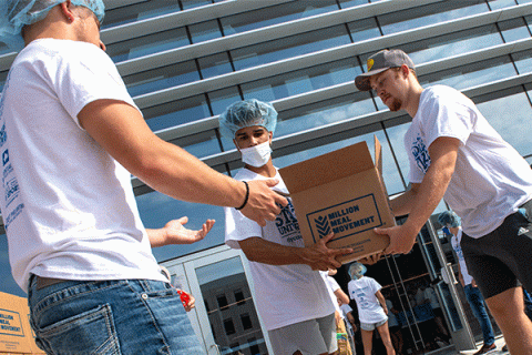 Three male students wearing informal outdoor clothing, including white Indiana State University t-shirts, and blue surgical caps, stand outside a University building with a glass front as they stack brown boxes containing food supplies for hungry families. The boxes have “Million Meal Movement” printed on the side. Other students can be seen working in the background. 