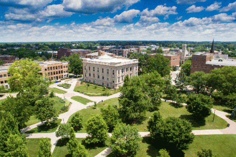 Elevated drone image of the Indiana State University campus, with Normal Hall, a white stone building, pictured in the center and other brick buildings visible nearby. The campus is shown in summer with green trees and grass visible in the foreground and around and behind the various buildings. 