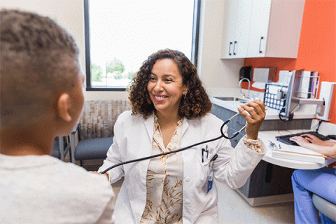 A dark-skinned middle- aged female wears a white coat over a white dress shirt with gold floral patterns. She holds a stethoscope while smiling and talking with a young patient with his back to the camera in a doctor’s office setting. A cabinet with a sink is visible behind the doctor. To the right, a person in blue scrubs is sitting and typing on a keyboard. 