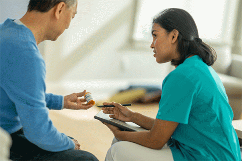 A senior gentleman of Asian descent, sits in a living room in his home with his nurse practitioner, during a home care visit. He wears a blue long-sleeved shirt and dark blue jeans. He holds a bottle of prescription pills. The practitioner is a tan-skinned woman with dark black hair pulled back behind her shoulders. She wears teal scrubs and khaki pants.  