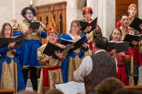 A group of singers in medieval-style costumes performs together. They hold music sheets and display various expressions of concentration and joy. The background features ornate wooden architecture, enhancing the festive atmosphere.