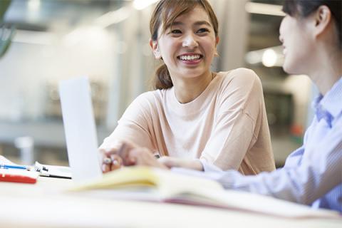 Two smiling female students of Asian heritage sit in a well-lit study area talking and laughing. The woman at left faces the camera and looks at her companion. She has medium-length brown hair in a ponytail and wears a light beige, long-sleeved shirt. Her friend has black hair and is looking away from the camera at the edge of the frame. She wears a blue-and-white pin-striped shirt, holds a pen, and has a laptop open before her. Books, pens, and notepads are visible but out of focus on a table.