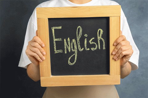 An individual in a white shirt, whose hands are visible and whose face is not, holds a square slate chalkboard framed in wood. The word ‘English’ is written on the chalkboard in large yellow letters.  