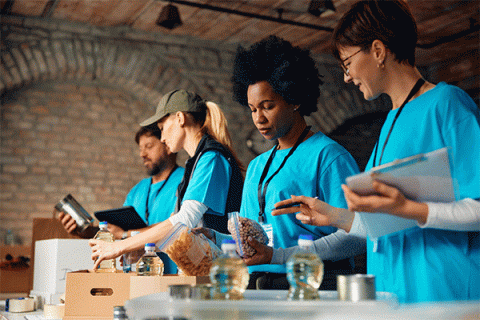 A diverse group of volunteers – three women and one man – work in a large room with brick walls to pack donated food items. They wear blue shirts and are placing Ziploc bags of food items, bottles of cooking oil, and cans of food into boxes. A smiling woman at the far right of the image holds a clipboard and appears to be directing the activity.