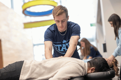 A male student with light brown hair, wearing a slate-blue t-shirt that says STATE on the front, presses down with both hands to massage the back of a man lying facedown on a massage table. The man on the table has short, dark hair and wears grey jeans and a beige shirt. In the background, slightly out of focus, two female students can be seen working with clients on other tables.   