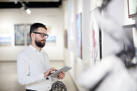 A white male with a dark brown beard, mustache, and glasses types into a tablet while looking at a piece of art on a gallery wall. He wears a white, long-sleeved, buttoned-up shirt. The artwork he is studying is not fully visible but appears to have splashes of magenta on it. Other pieces are visible on the walls behind him, and fragments of sculpture or 3D artworks are out of focus in the foreground.