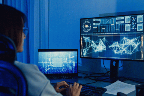 A woman with long dark hair and glasses sits facing two monitors, one a laptop and the other a desktop with a large screen, in a darkened room with blue lighting. The main monitor displays digital data, while the small monitor is partially visible. Neatly arranged papers lie on the desk.