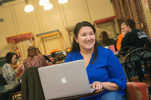 A female with black hair wearing a blue shirt sits at a table in a café, working on an Apple laptop. The background includes other patrons sitting at tables. The café features light fixtures, wooden furniture, and brick walls.