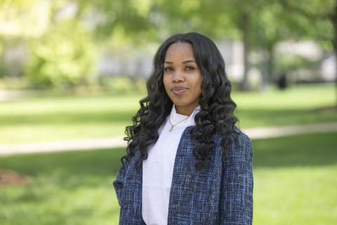 Shanisa Stinson stands smiling outdoors in a campus setting. She has long, curly black hair and is wearing a white shirt and a blue textured jacket. The background features green grass, trees, and blurred foliage.