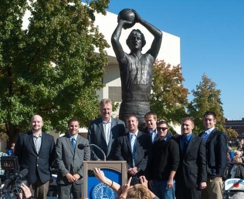Eight men stand smiling in front of a large bronze basketball statue at Indiana State University. One man speaks from a podium with the university seal, surrounded by trees and campus buildings on a sunny day.