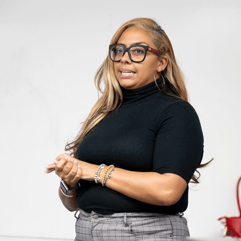 A Black woman with long highlighted hair speaks while standing. She is wearing large black glasses, a black turtleneck, hoop earrings, and patterned pants, with her hands clasped in front of her against a plain background.