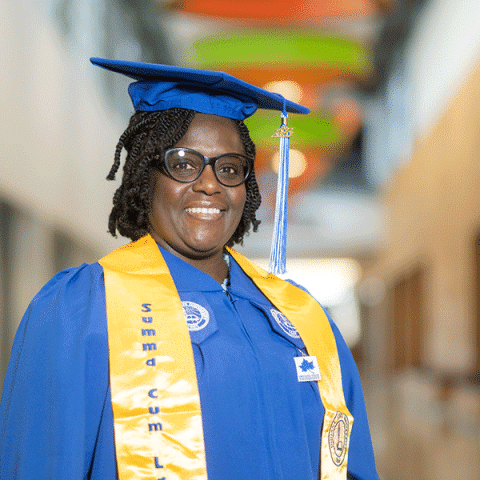 A Black woman wearing glasses and a blue graduation cap and gown smiles while standing indoors. She has a yellow stole that reads “Summa Cum Laude,” and a blue tassel hangs from her cap. Colorful ceiling panels are blurred in the background.