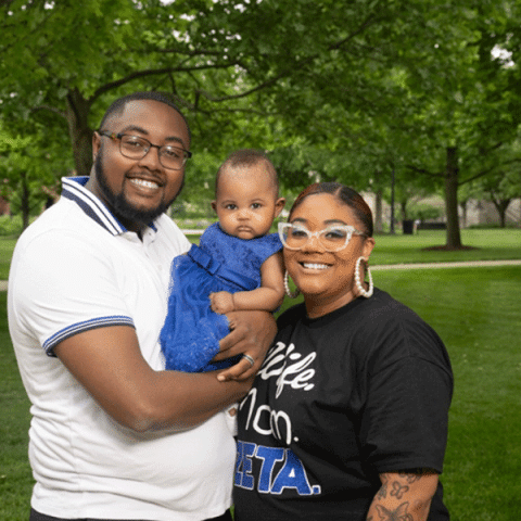 A Black man wearing glasses holds a baby in a blue dress while standing beside a smiling Black woman with clear-framed glasses. They are outdoors in a grassy park with trees in the background.