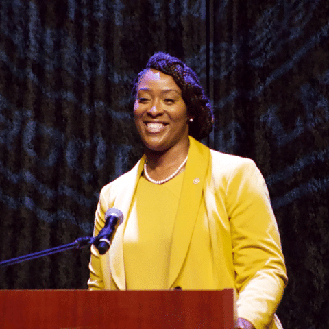 A Black woman with braided hair stands at a podium, smiling while speaking. She is wearing a yellow blazer and a pearl necklace. A microphone is positioned in front of her, and a dark curtain backdrop is behind her.