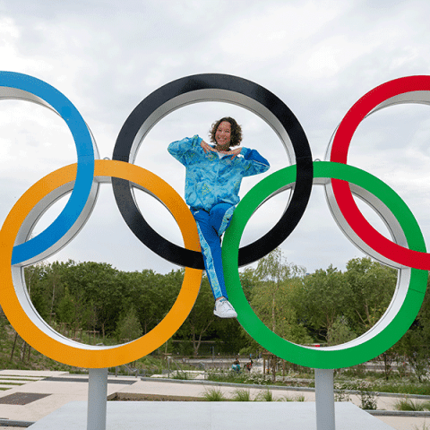 A woman wearing a bright blue athletic outfit sits inside the black Olympic ring of a large outdoor Olympic rings sculpture, smiling and posing with her hands. The other colored rings surround her, with trees and a cloudy sky in the background.