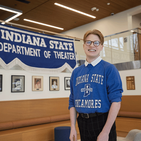 A white man wearing glasses and a blue “Indiana State Sycamores” sweatshirt stands indoors, smiling in front of a large banner that reads “Indiana State Department of Theater,” with framed photos displayed on the wall behind him.