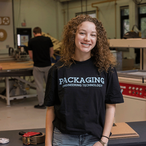A young woman with light skin and curly hair stands smiling in a workshop, wearing a black T-shirt that reads “Packaging Engineering Technology.” Work tables and equipment are visible in the background, with another person working behind her.