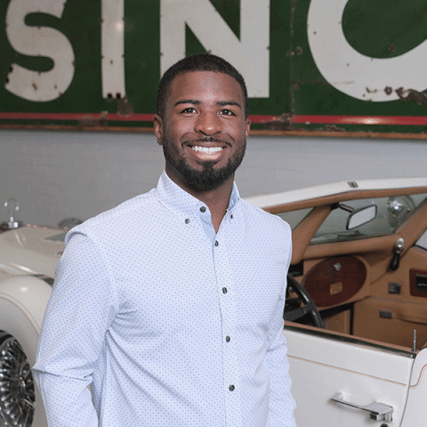A Black man wearing a light button-down shirt stands smiling indoors in front of a white vintage car, with a large, weathered sign hanging on the wall behind him.