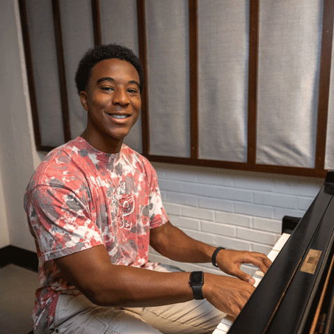 A young Black man with short natural hair, wearing a red and white tie-dye shirt and a smartwatch, sits at a piano smiling. The room has white brick walls and acoustic panels behind him.