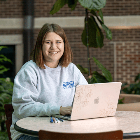 A white woman with straight shoulder-length light brown hair sits at a round table with a laptop in front of her. She is smiling and wearing a light gray sweatshirt that says “Honors College.” Pens are on the table, and brick walls and plants are visible in the background.
