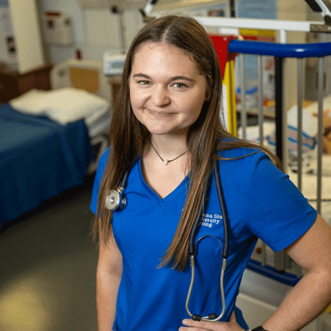 A young white woman with long straight brown hair stands smiling in a nursing skills lab. She is wearing a blue scrub top with a stethoscope around her neck. A hospital bed and medical equipment are visible in the background.