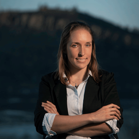 A white woman with light brown hair wearing a black blazer over a light blue striped shirt stands outdoors with her arms crossed, smiling slightly. The background is blurred, showing a dark building landscape at dusk.