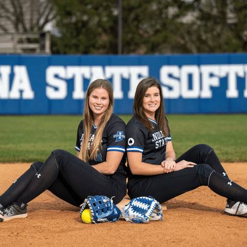 Two white women in black Indiana State softball uniforms sit back-to-back on the infield dirt, smiling. Each has a softball glove in front of her, and a blue “INDIANA STATE SOFTBALL” outfield wall is visible behind them.