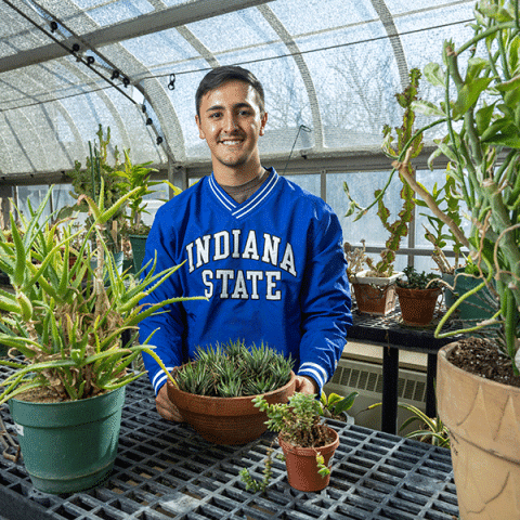 A young man wearing a blue “Indiana State” sweatshirt stands smiling in a greenhouse, holding a potted plant among several other potted succulents and greenery.