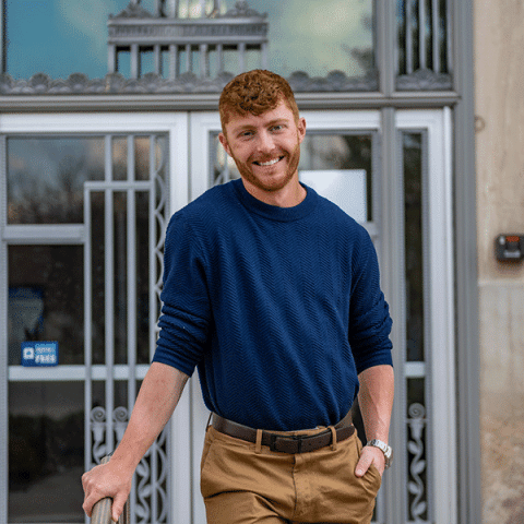 A white man with short curly hair and a trimmed beard, wearing a blue sweater and khaki pants, stands smiling outdoors with one hand resting on a railing in front of a building with decorative glass and metal doors.