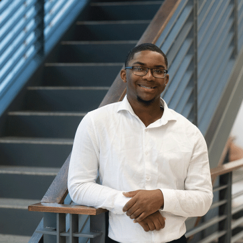 A Black man with short hair and glasses stands smiling beside a staircase railing. He is wearing a white button-down shirt and has his hands lightly crossed in front of him.