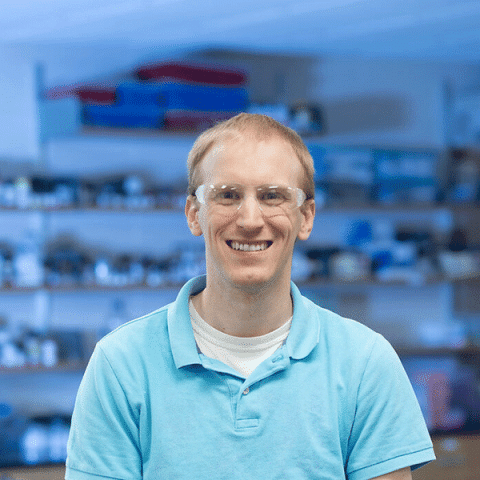 A white man wearing safety glasses and a light blue polo shirt stands smiling in a lab space, with shelves of tools and supplies in the background.