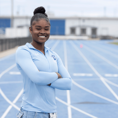 A Black woman wearing a light blue athletic top stands smiling with her arms crossed on a blue running track, with stadium buildings visible in the background.