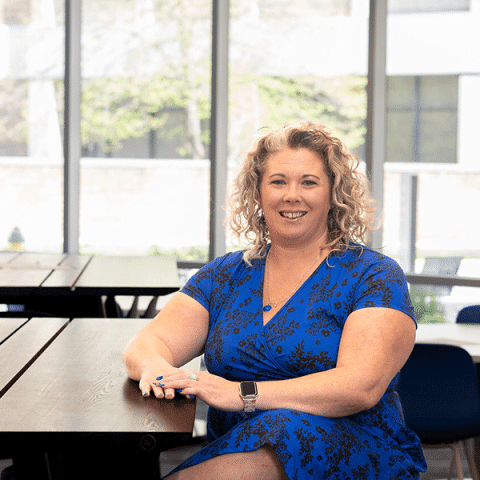 A white woman with curly blonde hair sits at a table, smiling. She is wearing a blue patterned dress and a smartwatch. Large windows behind her show trees and a bright outdoor scene.