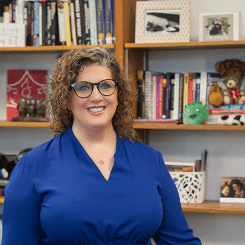 A white woman with curly light brown hair stands smiling in front of bookshelves. She is wearing glasses and a bright blue blouse. Books, framed photos, and small decorative items are visible behind her.